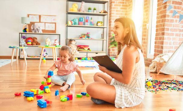 Beautiful psycologist and blond toddler girl doing therapy building tower using plastic blocks at kindergarten - Photo, Image