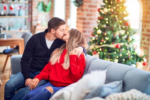 Young beautiful couple smiling happy and confident. Sitting on the sofa hugging and kissing around christmas tree at home - Photo, Image