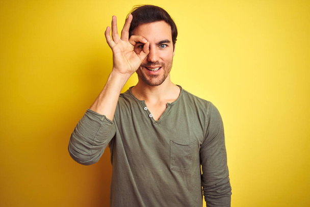 Młody przystojny mężczyzna ubrany w luźny t-shirt stojący nad odizolowanym żółtym tłem wykonujący ok gest z uśmiechem dłoni, okiem patrzącym przez palce ze szczęśliwą twarzą. - Zdjęcie, obraz