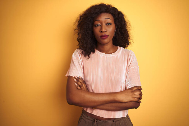 Afroamerykanka ubrana w różowy t-shirt stojąca nad odizolowanym żółtym tłem sceptyczna i nerwowa, dezaprobata wyraz twarzy z skrzyżowanymi ramionami. Osoba negatywna. - Zdjęcie, obraz