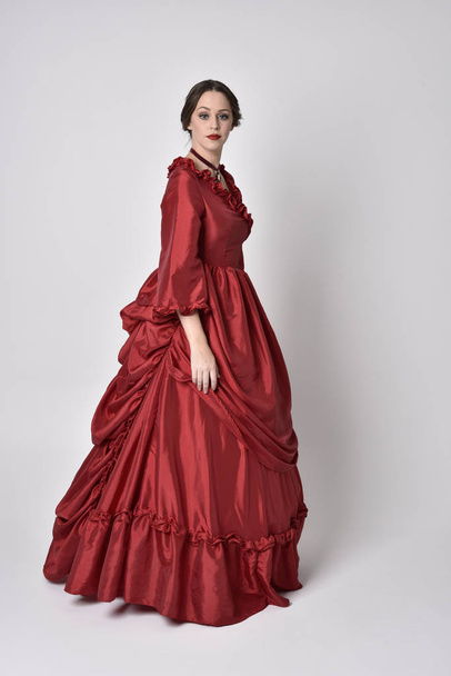 портрет брюнетки в красном шелковом викторианском платье. Стоящая поза на белом студийном фоне
. - Фото, изображение