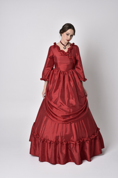 full length portret van een brunette meisje in een rode zijden victoriaanse jurk. Staande houding op een witte studio achtergrond. - Foto, afbeelding