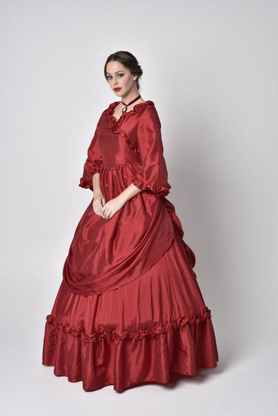 full length portrait of a brunette girl wearing a red silk victorian gown. Standing pose on a white studio background. - Photo, Image