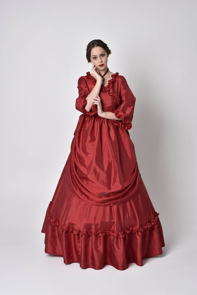 портрет дівчини брюнетки в червоному шовковому вікторіанському сукні. Позування на білому фоні студії
. - Фото, зображення