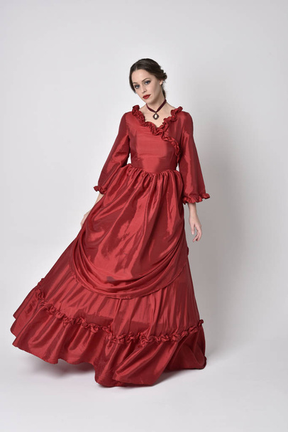 Täyspitkä muotokuva brunette tyttö yllään punainen silkki viktoriaaninen puku. Seisova poseeraa valkoisella studiolla taustalla
. - Valokuva, kuva