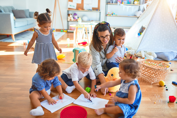 Красивий вчитель і група малюків, які сидять на підлозі малюють за допомогою паперу і олівця навколо безлічі іграшок в дитячому саду
 - Фото, зображення