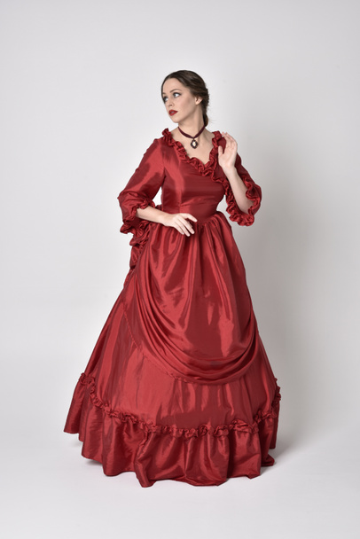 pełnowymiarowy portret brunetki w czerwonej jedwabnej wiktoriańskiej sukni. Pozycja stojąca na białym tle studia. - Zdjęcie, obraz