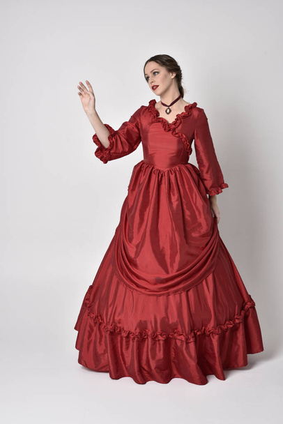 full length portrait of a brunette girl wearing a red silk victorian gown. Standing pose on a white studio background. - Zdjęcie, obraz