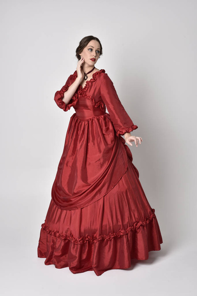full length portret van een brunette meisje in een rode zijden victoriaanse jurk. Staande houding op een witte studio achtergrond. - Foto, afbeelding