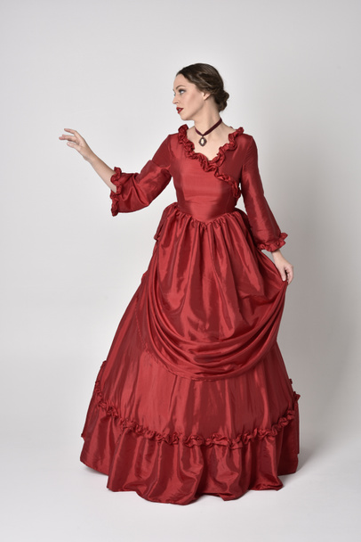 full length portrait of a brunette girl wearing a red silk victorian gown. Standing pose on a white studio background. - Фото, зображення