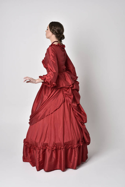 full length portrait of a brunette girl wearing a red silk victorian gown. Standing with back to the camera on a white studio background. - Photo, Image