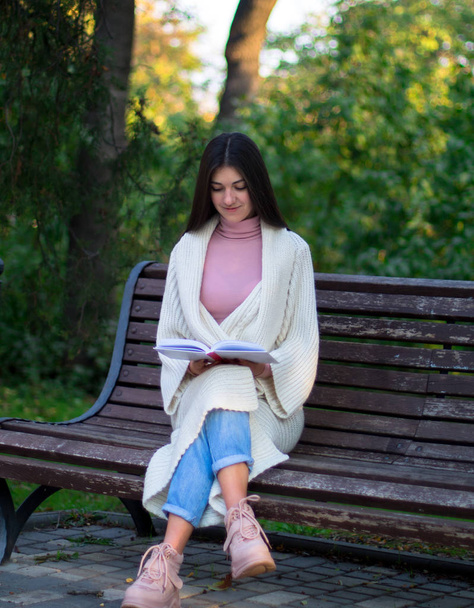 Ένα κορίτσι με άσπρη ζακέτα διαβάζει ένα βιβλίο στο παγκάκι στο πάρκο. - Φωτογραφία, εικόνα