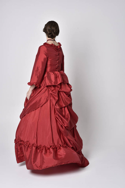 full length portrait of a brunette girl wearing a red silk victorian gown. Standing with back to the camera on a white studio background. - Foto, Bild