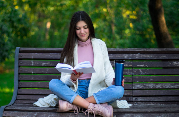 молода жінка читає книгу на лавці в парку, термо-кухоль на лавці
 - Фото, зображення