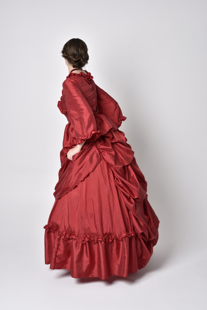 full length portrait of a brunette girl wearing a red silk victorian gown. Standing with back to the camera on a white studio background. - 写真・画像
