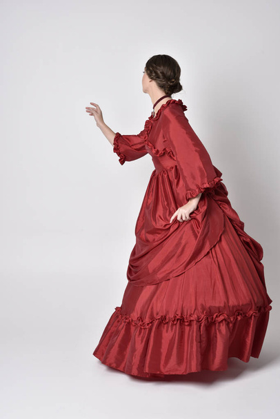 портрет брюнетки в красном шелковом викторианском платье. Стоя спиной к камере на белом фоне студии
. - Фото, изображение