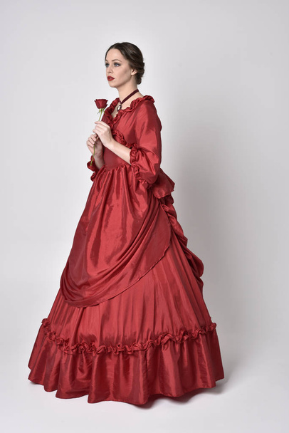  portrét brunetky v červených hedvábných viktoriánských šatech. Stojící se zády ke kameře na bílém pozadí studia. - Fotografie, Obrázek