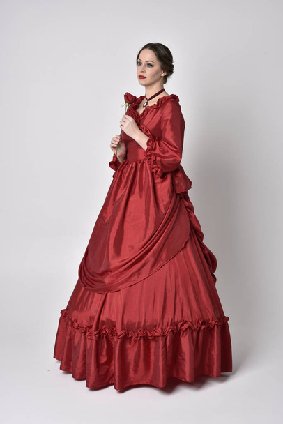  retrato de una chica morena con un vestido victoriano de seda roja. De pie con la espalda a la cámara en un fondo de estudio blanco
. - Foto, Imagen