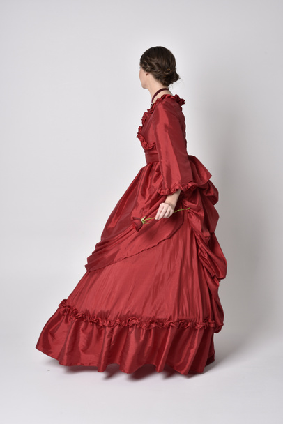  portrait of a brunette girl wearing a red silk victorian gown. Standing with back to the camera on a white studio background. - Fotografie, Obrázek