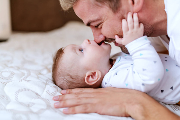 Παιχνιδιάρικο, αξιολάτρευτο καυκάσιο αγοράκι έξι μηνών ξαπλωμένο στο κρεβάτι και δαγκώνει τη μύτη του μπαμπά του. Ο πατέρας αγκαλιάζει το μωρό και γελάει. - Φωτογραφία, εικόνα