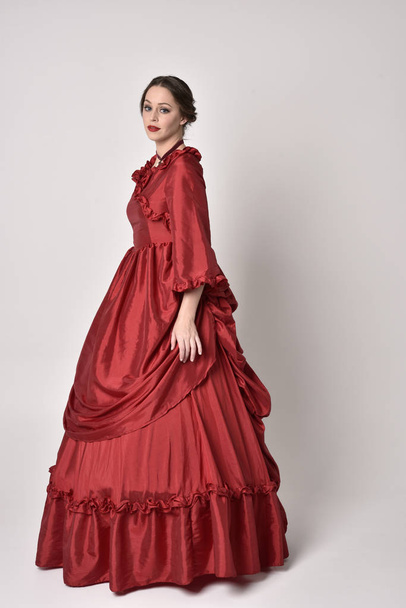 Kırmızı ipek Victoria elbisesi giyen esmer bir kızın tam boy portresi. Beyaz bir stüdyonun arka planında kameraya karşı durmak.. - Fotoğraf, Görsel