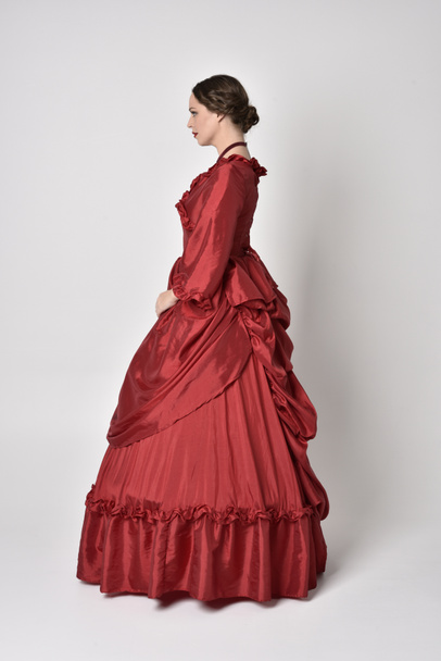 full length portret van een brunette meisje in een rode zijden victoriaanse jurk. Staande met rug naar de camera op een witte studio achtergrond. - Foto, afbeelding