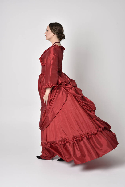 full length portrait of a brunette girl wearing a red silk victorian gown. Standing with back to the camera on a white studio background. - Fotografie, Obrázek