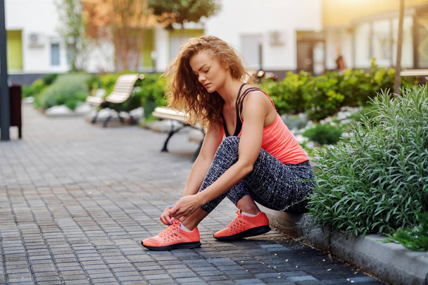 Charming fit caucasian woman dressed in sports wear sitting on roadside and tying shoelace. Park exterior. - Photo, Image