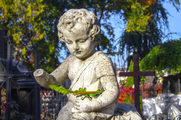 Estatua decapitada de niño arrodillado ángel con los brazos cruzados contra el cielo azul y lápida del cementerio con fondo de crucifijo
 - Foto, Imagen