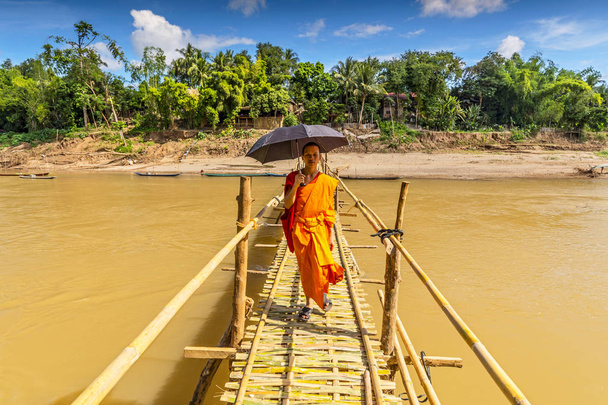 Молодой буддийский монах проходит по бамбуковому мосту через Нам-хан в Луангпрабанг-Лаосе
. - Фото, изображение