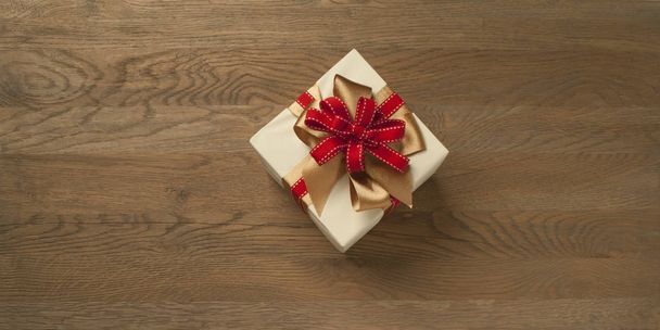 Рождественская подарочная коробка, завязанная красным и золотым бантом над деревянным столом. Горизонтальная композиция с копировальным пространством слева и справа от коробки
. - Фото, изображение