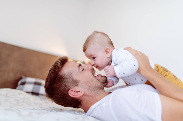 Guapo padre joven acostado en la cama en el dormitorio y levantando a su amoroso niño riendo de seis meses de edad. Concepto de amor incondicional
. - Foto, Imagen