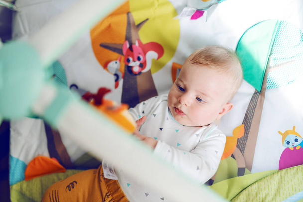 To view of curious baby boy six months old dressed in bodysuit lying in bed and playing with his crib toys. - 写真・画像
