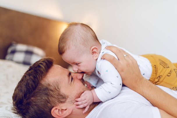 Sorrindo caucasiano bonito pai deitado na cama no quarto e segurando seu amoroso adorável filho de seis meses de idade em seu peito. Um bebé a morder o nariz do pai. Conceito de amor incondicional
. - Foto, Imagem