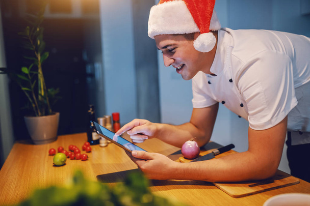 Handsome caucasian smiling chef in white uniform and with santa hat on head using tablet while leaning on kitchen counter. On kitchen counter are cherry tomatoes, avocado, spices and onion. - Photo, Image