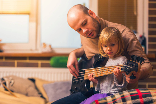 Cute little girl i jej ojciec grają na gitarze basowej i uśmiecha siedząc na kanapie w domu. Spędzać razem czas, uczyć się grać na gitarze - Zdjęcie, obraz
