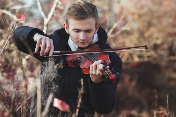 πορτρέτο ενός νεαρού κομψού βιολιστή που παίζει βιολί στο χώρο του φθινοπώρου στο γρασίδι, νεαρός άνδρας που ασχολείται με τη μουσική τέχνη, έννοια της κλασικής μουσικής, χόμπι και να χαλαρώσετε - Φωτογραφία, εικόνα