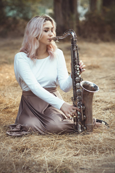 portrait of a beautiful young woman sitting on the dry grass leanher head on saxophone, romantic blonde girl close eyes and relaxing - Φωτογραφία, εικόνα
