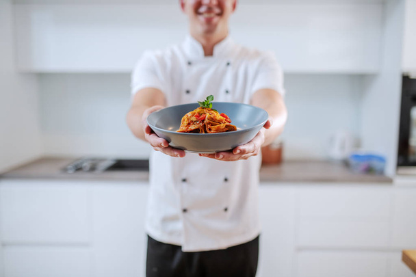 Усміхнений кавказький креативний шеф-кухар у білій формі тримає тарілку з тальятеллю, стоячи на кухні. Вибірковий фокус на тарілці
. - Фото, зображення