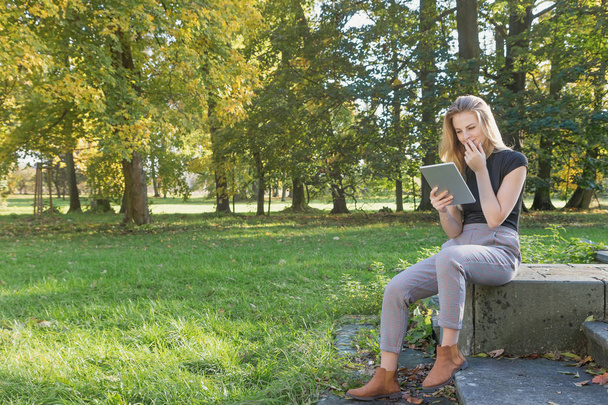 Smiling young girl is sitting in the park looking at the tablet - Photo, Image