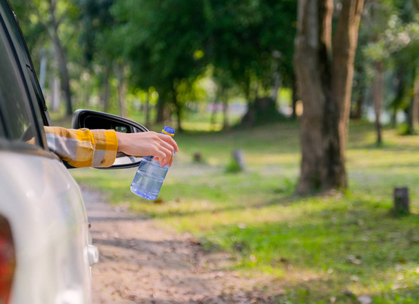 Kobieta trzyma niebieską butelkę przez okno samochodu i wygląda na to, że wyrzuci butelkę na bok jadąc z myślą o bezpiecznym środowisku z ludzkich śmieci.. - Zdjęcie, obraz