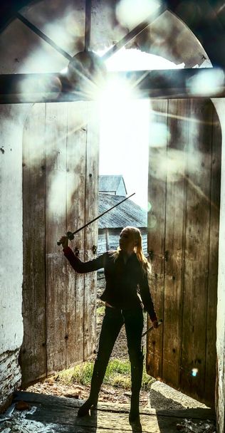 mulher loira guerreira com uma espada em pé no velho, retro, vintage, velho fundo da porta aberta. elfo feminino medieval com espada olhando dentro da igreja
 - Foto, Imagem