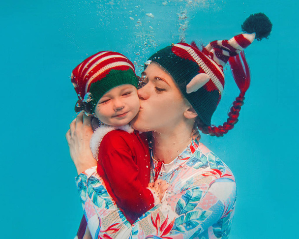Mujer caucásica (madre) besando a su hijo (bebé) bajo el agua en año nuevo y trajes de elfo de Navidad en la piscina
 - Foto, Imagen