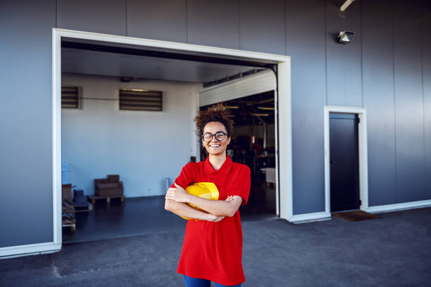 Successful Caucasian smiling female employee standing in front of printing shop with arms crossed and holding helmet. - Photo, Image