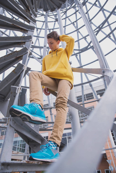 A young guy in a yellow hoodie and turquoise sneakers stands on a spiral staircase. Teen jumps off with a beautiful spiral staircase in the beautiful architecture - Foto, Imagem