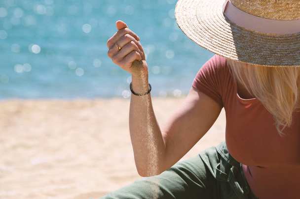 Woman with casual outfit and straw hat sitting on sandy beach against blue sea and sand flowing through her hands. Summer vacation concept - Φωτογραφία, εικόνα