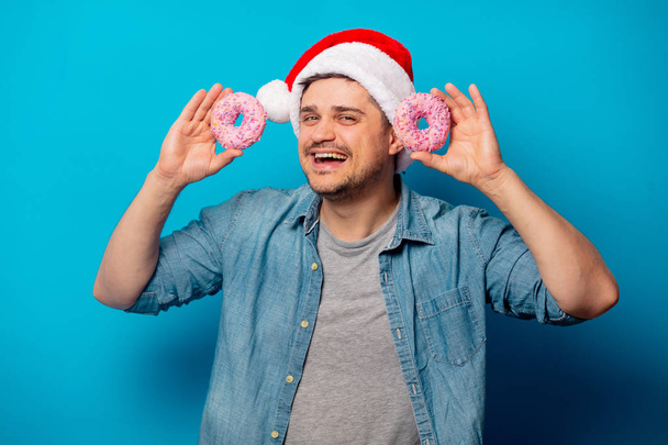 Όμορφος άντρας με Χριστουγεννιάτικο καπέλο με ντόνατς σε μπλε φόντο. - Φωτογραφία, εικόνα