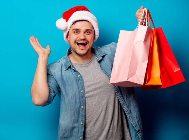 Handsome man in Christmas hat with shopping bags on blue backgro - Фото, изображение