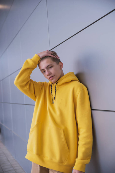 A serious young guy in a yellow hoodie, a cute stylish fashion man in bright clothes against a gray wall - Foto, afbeelding