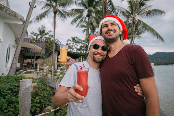 Молодая пара геев с коктейлями в рождественских шапках на океане
 - Фото, изображение